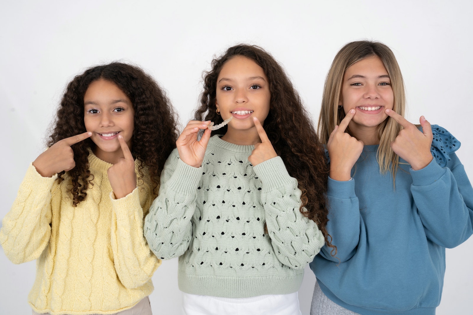 Three middle-school-aged girls point at their smiles. The girl in the middle holds up clear aligners.