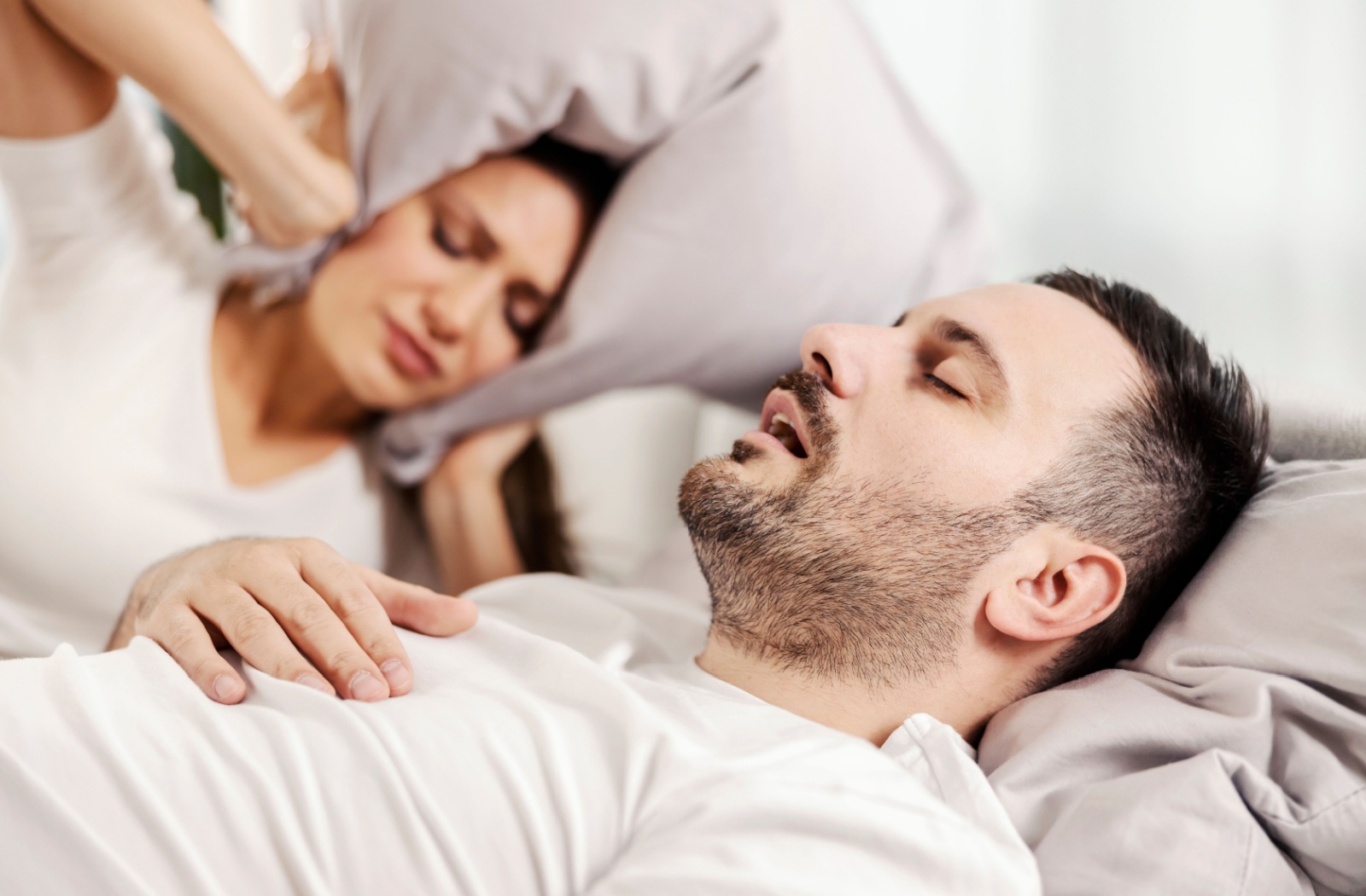 Two people lying in bed, one is snoring and the other is covering their ears with a pillow