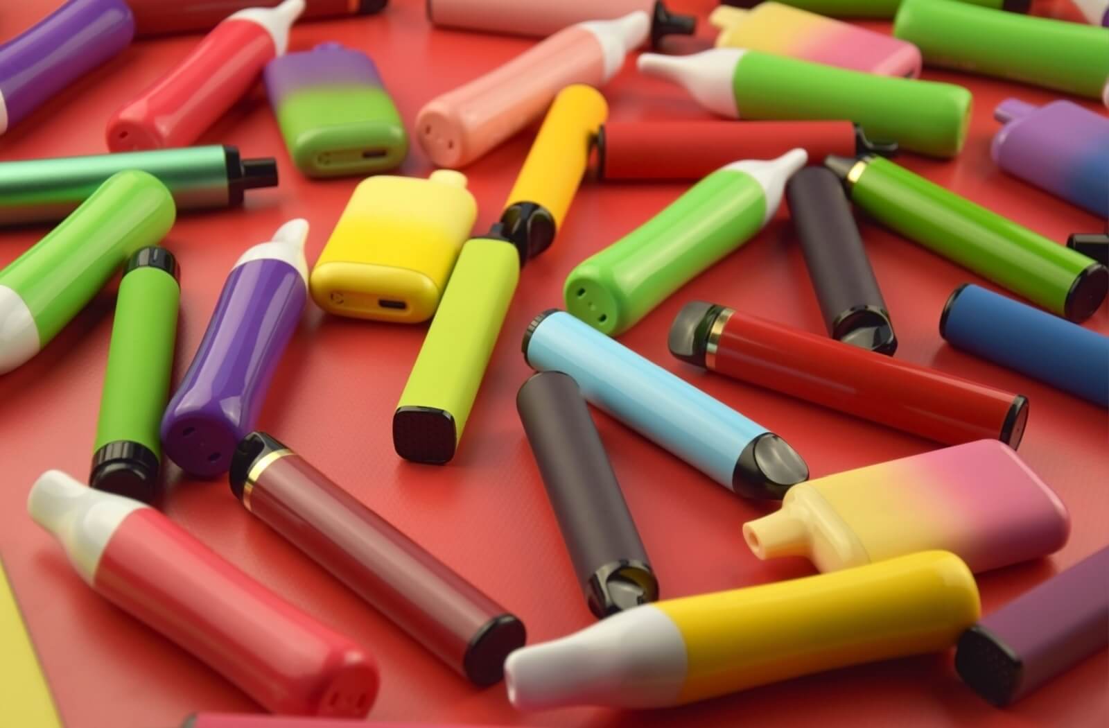 A large assortment of colorful disposable vaping devices scattered across a solid red surface.