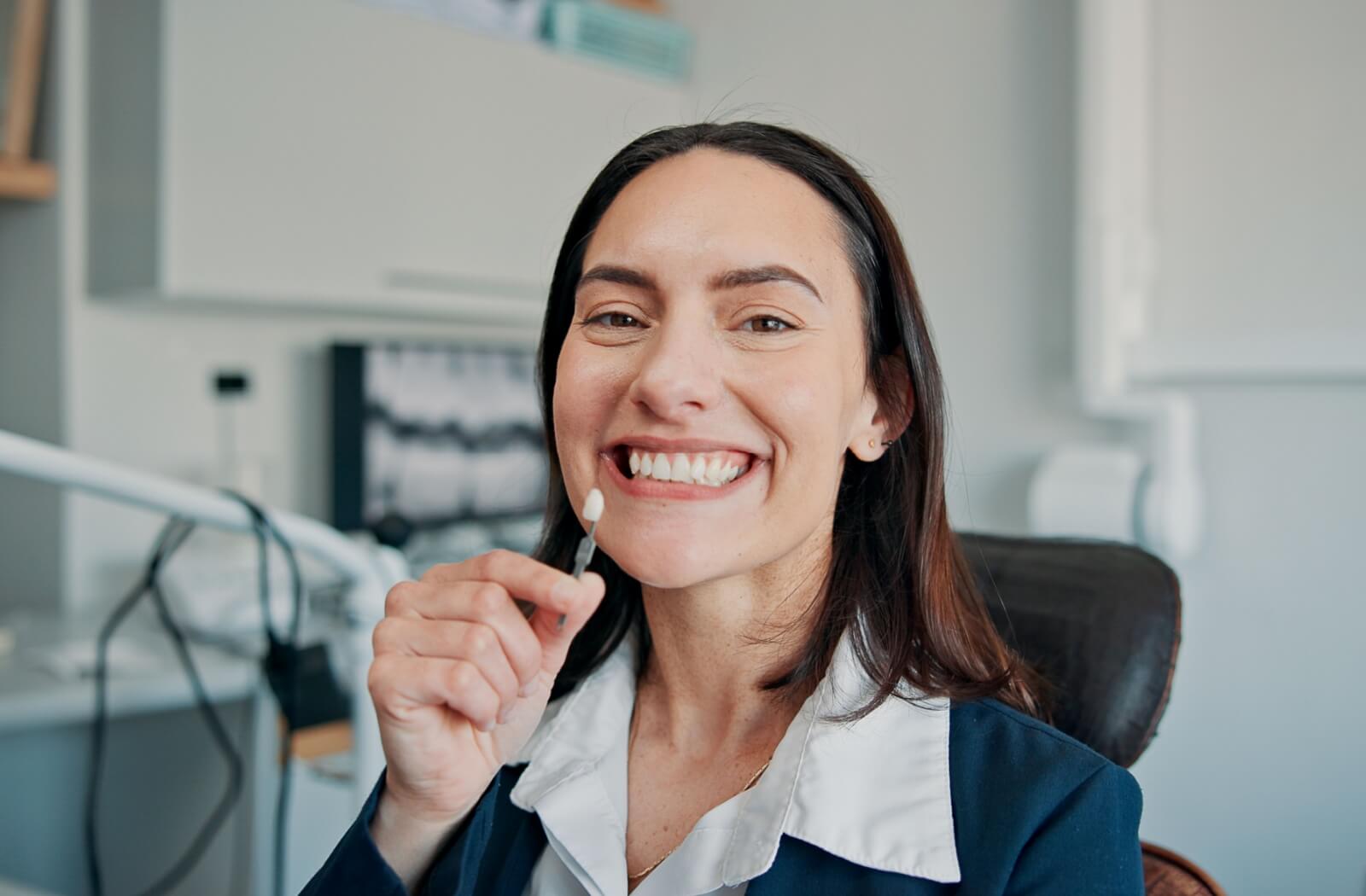 A smiling individual holding a tooth shade guide up to their teeth during a dental implant consultation.