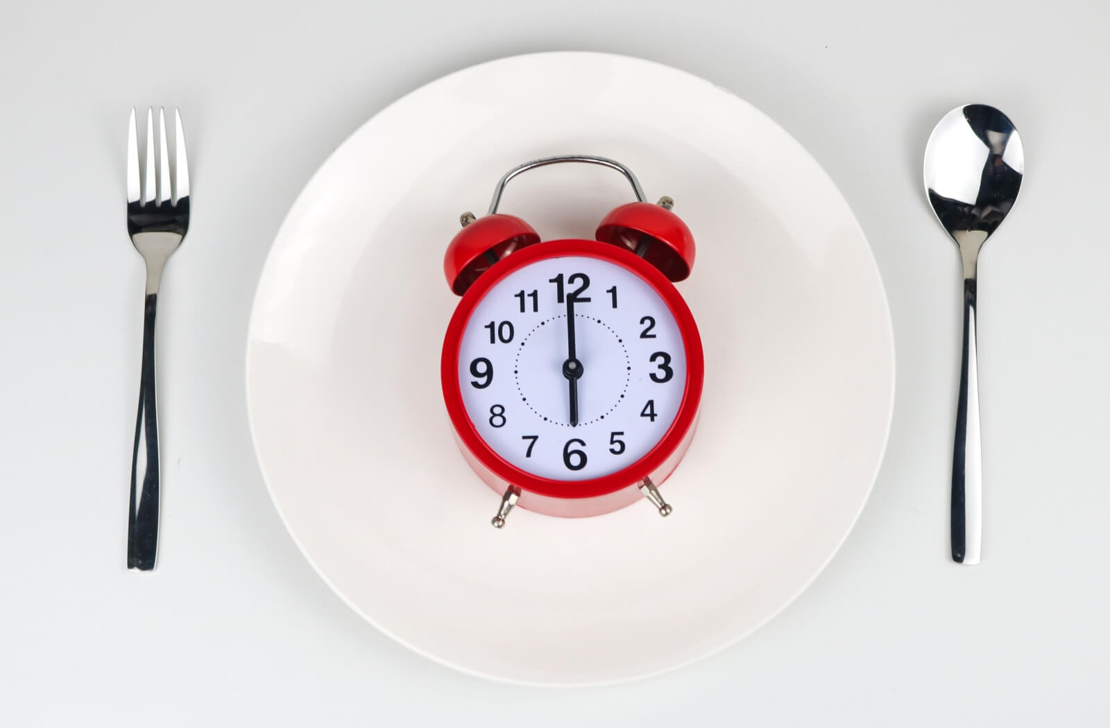 A top-down view of a red alarm clock sitting on a white dinner plate between a silver fork and spoon, symbolizing dental timelines.