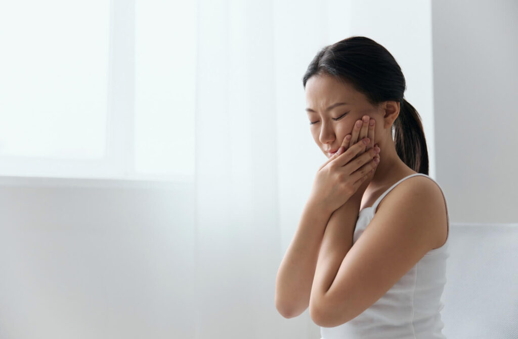A young woman sitting in a bright room, holding her jaw in pain to illustrate symptoms of wisdom teeth coming in.